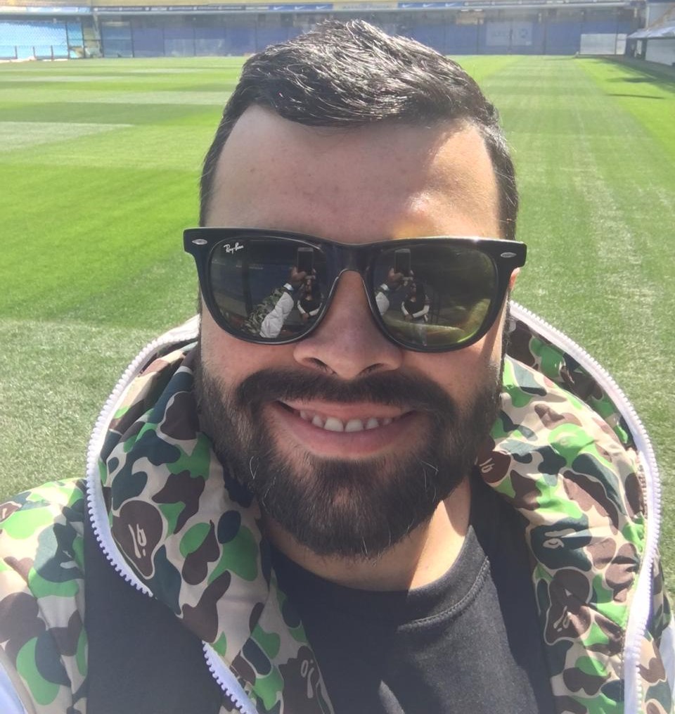 Bruno Tiago de jaqueta com estampa de camuflagem e zíper frontal e camiseta preta, em um campo de futebol bem cuidado com grama verde e linhas marcadas, de óculos escuro e sorrindo.